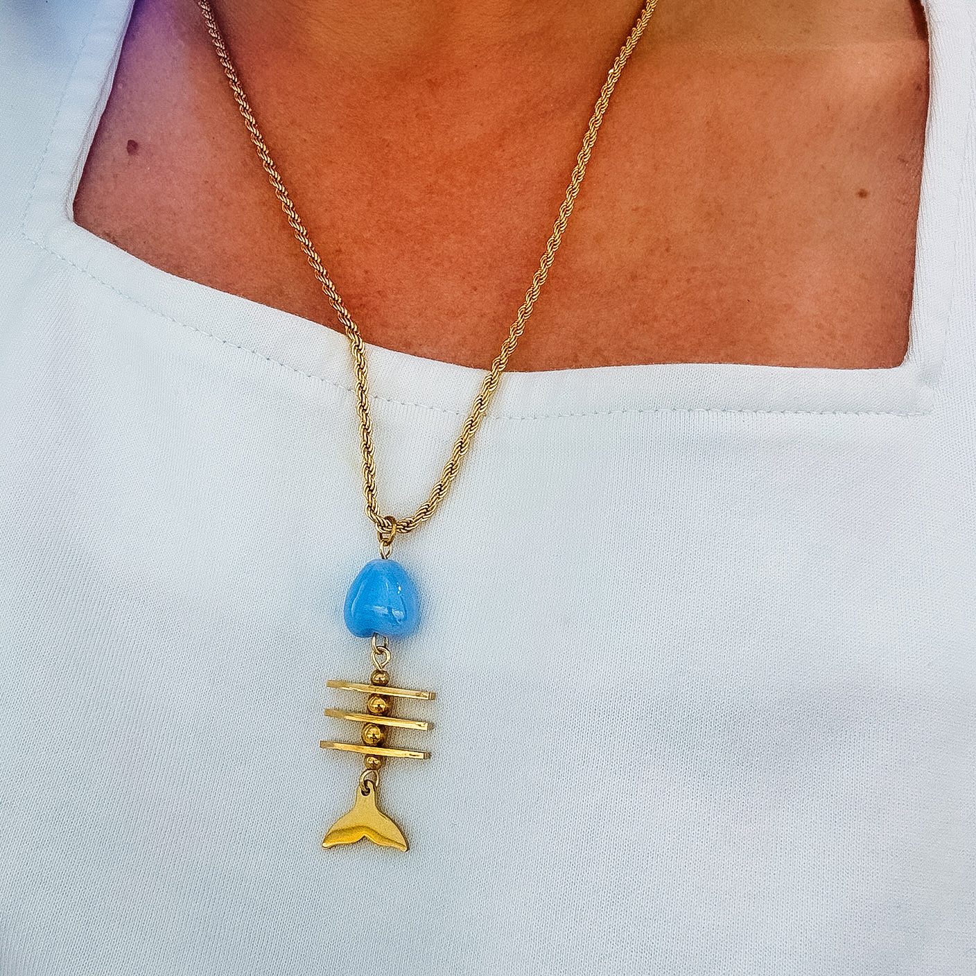 Dainty gold layered necklace featuring a twisted rope chain with a playful pendant made of a blue fish-shaped bead, gold bar details resembling a fish skeleton, and a gold tail charm, waterproof, hypoallergenic, tarnish free, statement accessories, wholesale jewelry for women boutiques